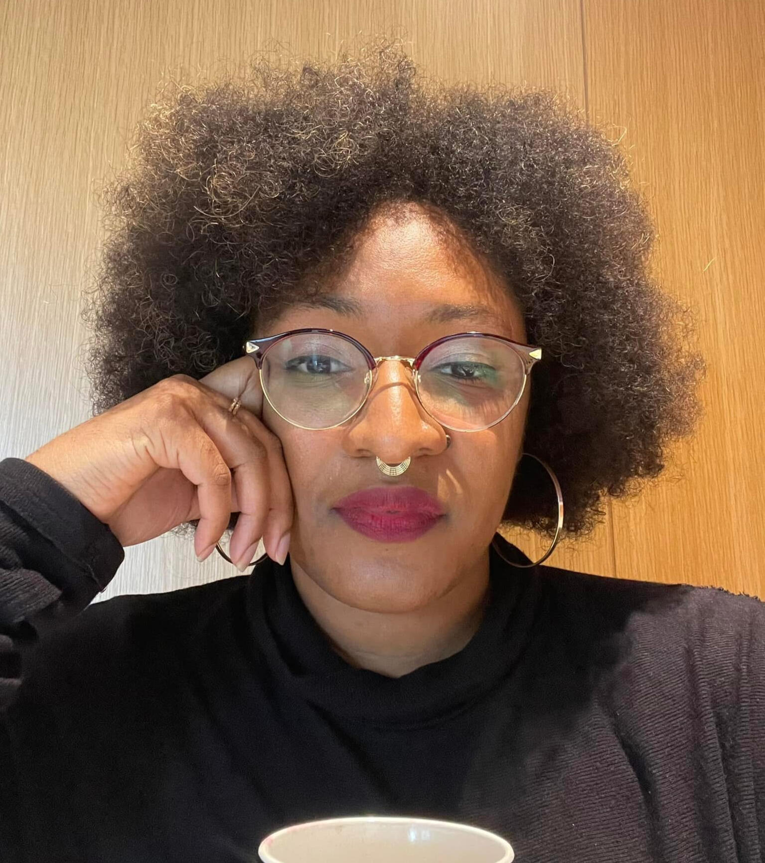 AI’s Out Here SPEEDRUNNING. 😲 Let Me Put You On. Joya Jones, a Black woman with a voluminous afro, gold-rimmed glasses, and a septum ring, sits against a warm wood background. In a black turtleneck and gold hoops, she rests her head on her hand, radiating confidence, deep knowledge, and AI mastery. 💡✨
